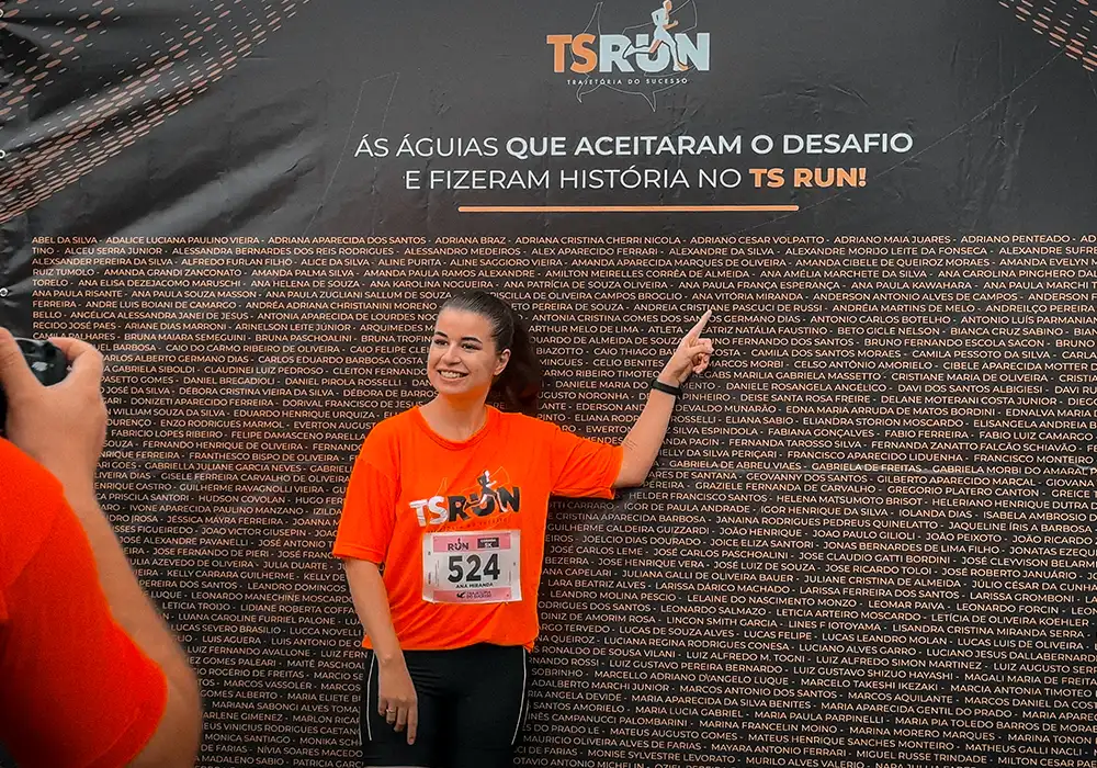 mulher tirando foto na frente de um banner do TS run com o nome das pessoas que fizeram parte